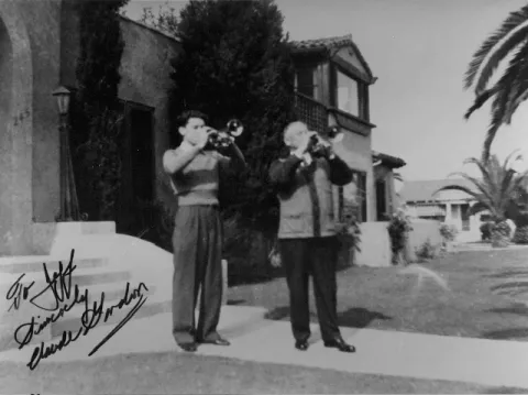 Two men playing trumpets outside a house, surrounded by palm trees.
