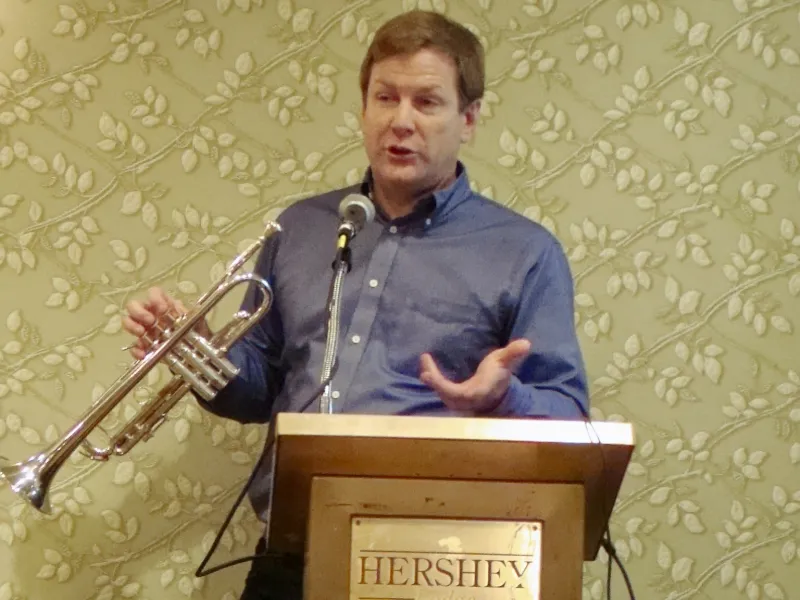 Jeff Purtle in a blue shirt speaks at a podium with a trumpet at the International Trumpet Guild's 2017 Convention in Hershey, Pennsylvania.